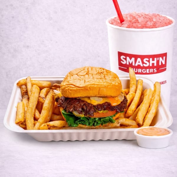 Smash'n Burgers cheeseburger with lettuce and sauce, surrounded by fries, dipping sauce, and a red frozen drink.